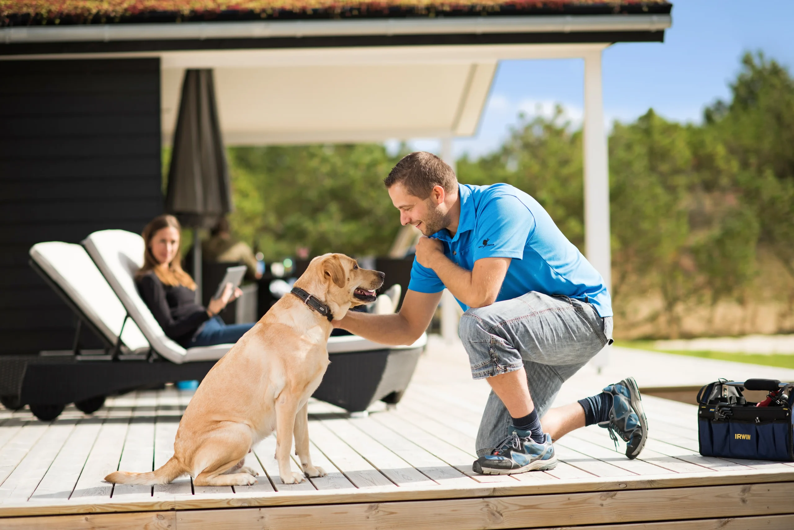 Esmark - Mann streichelt Hund. Frau liegt im Hintergrund auf der Terrasse